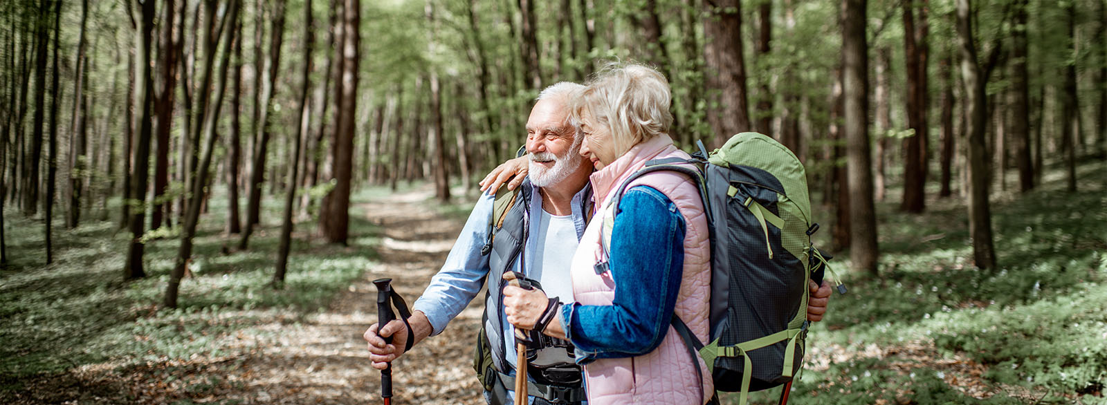 Senior couple hugging in the forest while hiking with backpacks and trekking sticks.