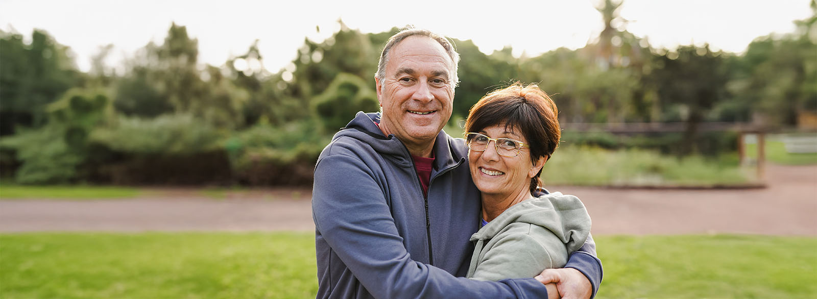 Happy senior couple hugging each other at city park while smiling on camera.