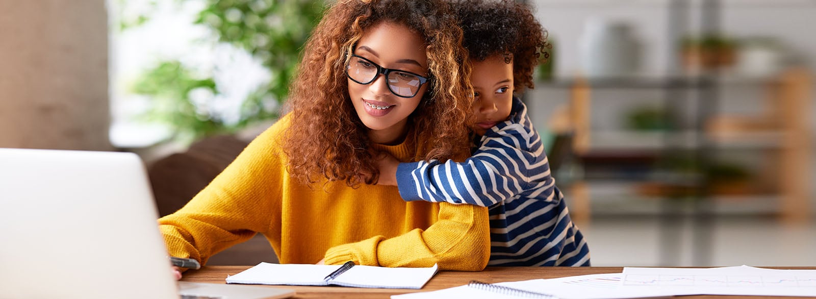 Young focused woman mother wearing eyeglasses using laptop and thinking about work task while small boy son gently hugs her.