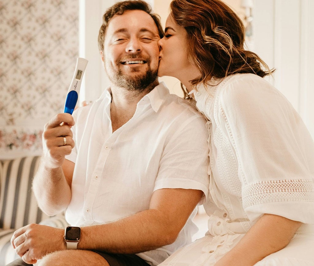 Woman kissing man holding a pregnancy test kit.